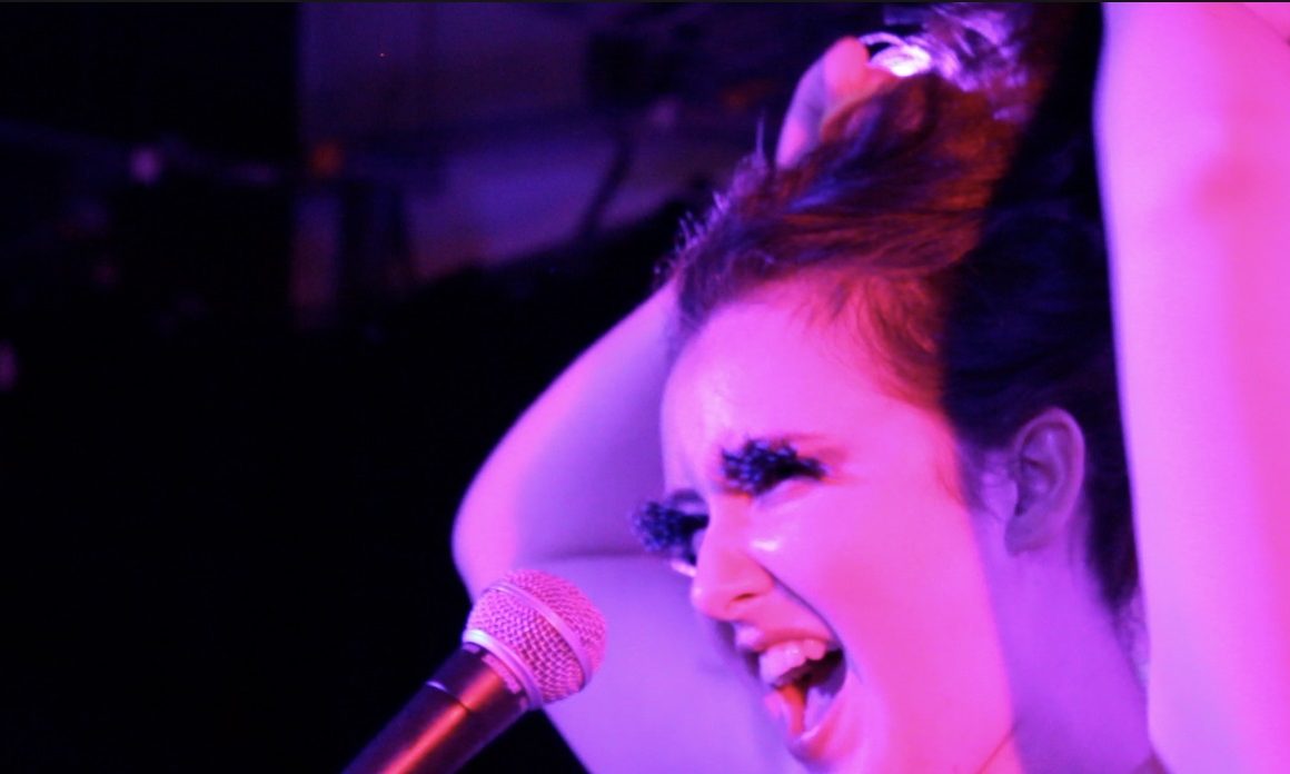 colour photograph, interior. White non-binary person is bathed in pink light both of their arms are above their head holding up their hair. They are singing into a microphone and have false lashes on.