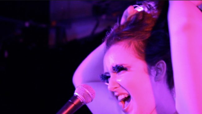 colour photograph, interior. White non-binary person is bathed in pink light both of their arms are above their head holding up their hair. They are singing into a microphone and have false lashes on.