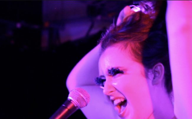 colour photograph, interior. White non-binary person is bathed in pink light both of their arms are above their head holding up their hair. They are singing into a microphone and have false lashes on.