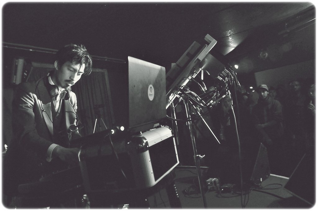 Black and white photo, interior. Human with black hair and olive skin in the left foreground, playing electric drums, electric keyboard and laptop on a stage, with headphones around their neck. A small crowd of humans standing listening to them to the right in the background.)